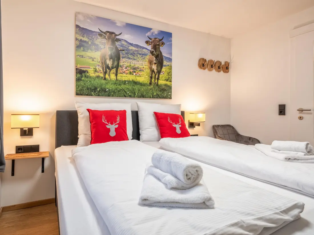 Een slaapkamer met een tweepersoonsbed, rode kussens met een hert en een grote foto van twee koeien in een weide.