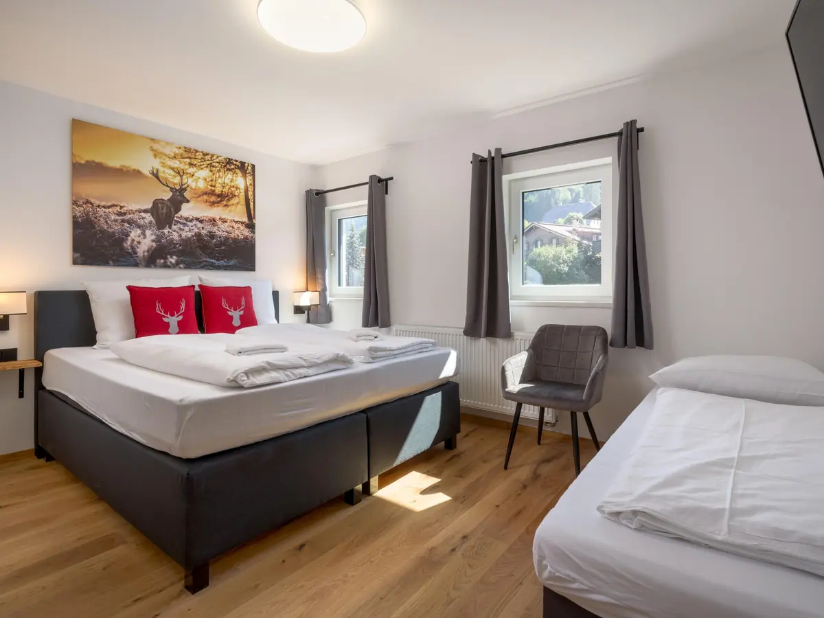 Ein Schlafzimmer mit Doppelbett, roten Kissen mit Hirschmotiv, einem Einzelbett und zwei Fenstern.