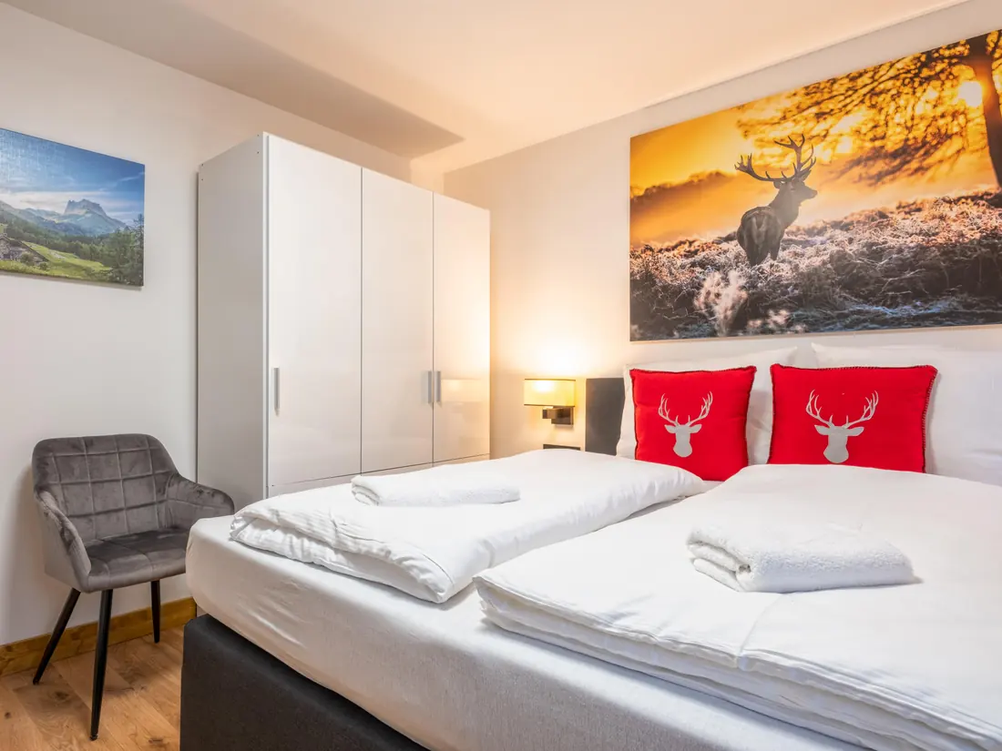 Een slaapkamer met een tweepersoonsbed, rode kussens met een hert en een witte kledingkast.