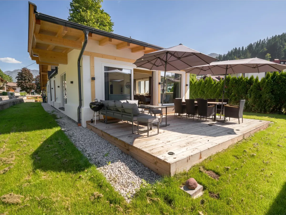 Die Holzterrasse im Sommer mit zwei Sonnenschirmen, Esstisch, Loungesofa und Grill, rundherum Rasen.