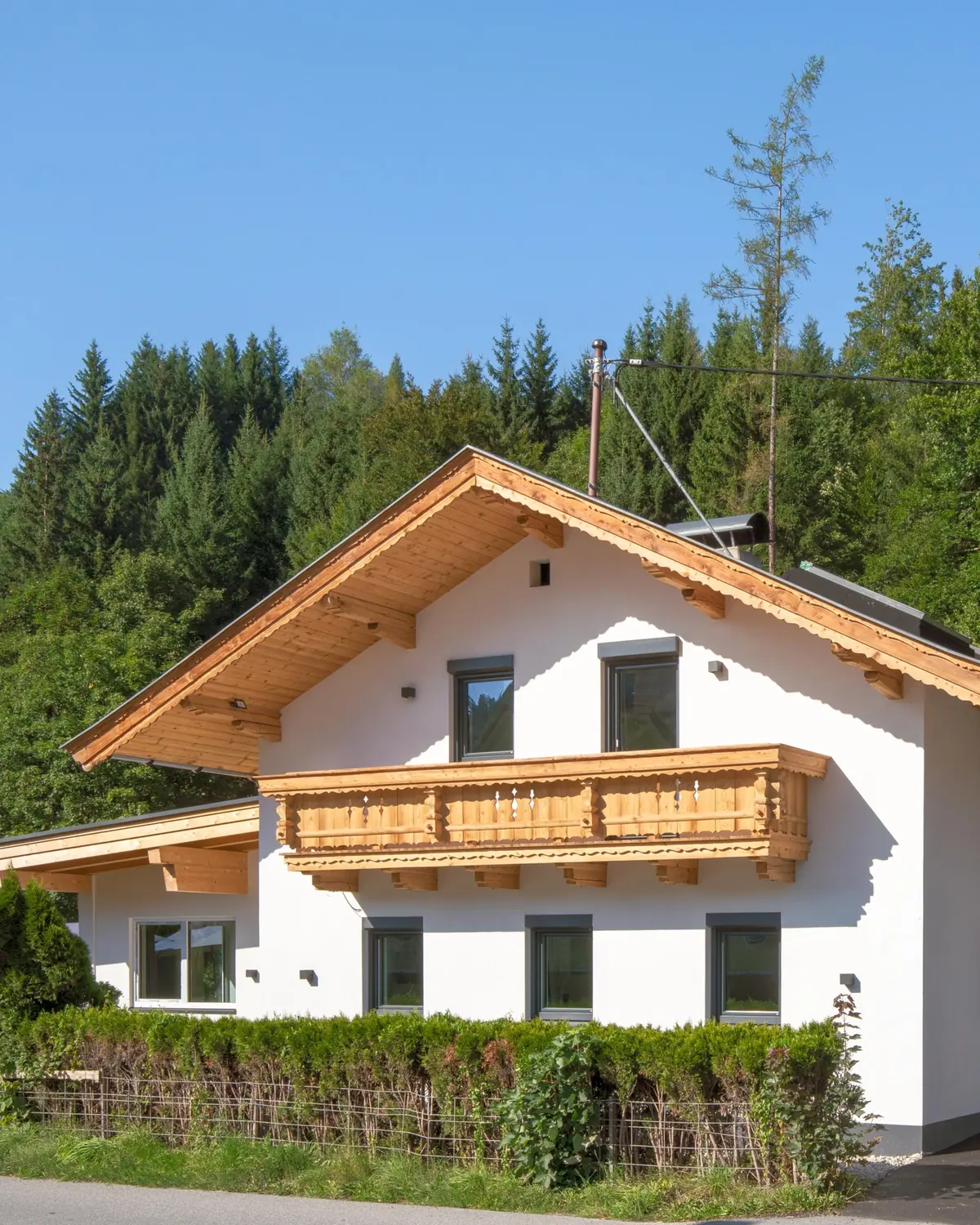 Chalet Hornblick vor grünem Wald: ein weißes Haus mit Holzgiebel, geschnitztem Holzbalkon und einer Hecke davor.