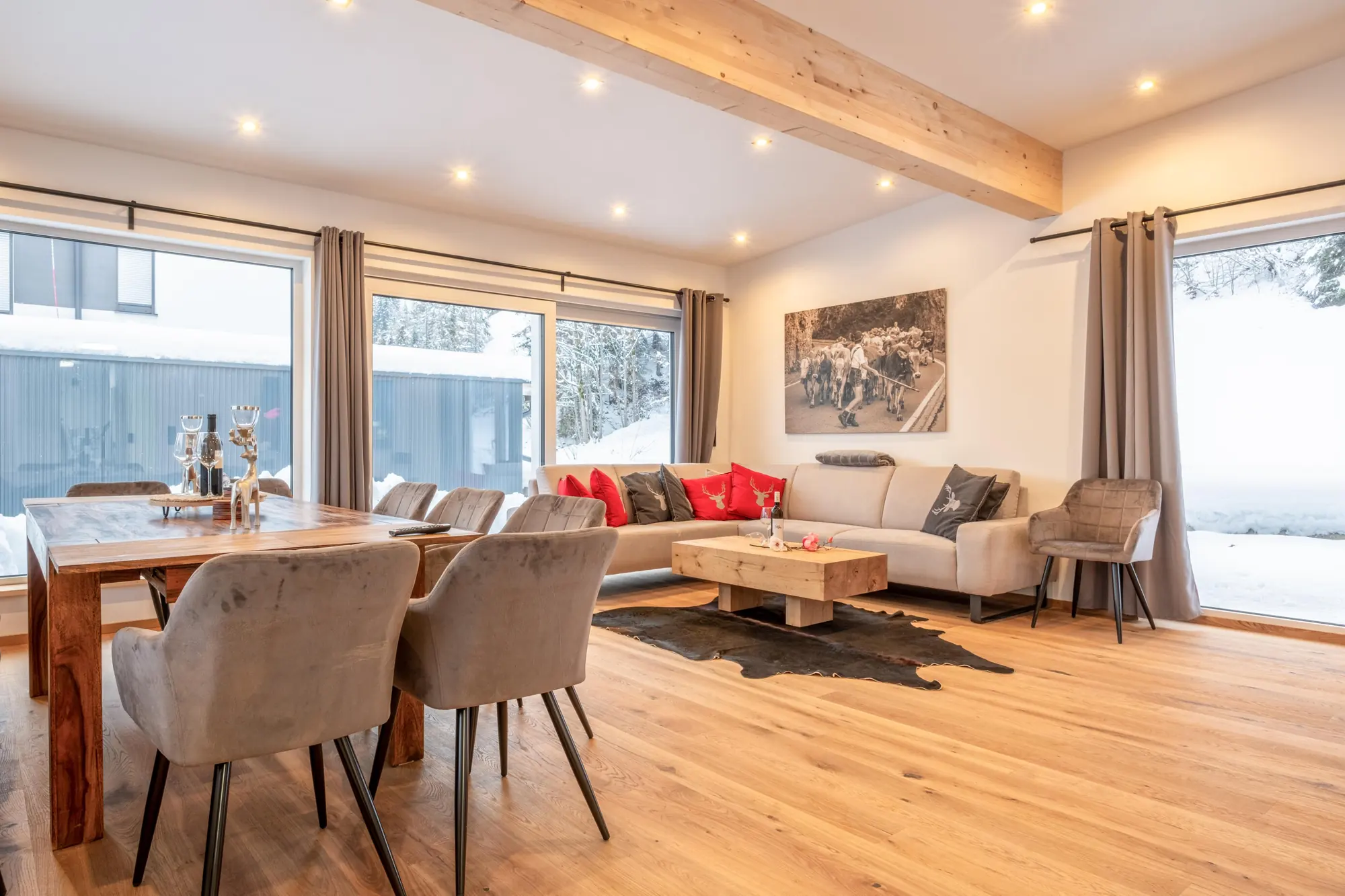 De woon- en eetkamer met eiken vloer, een houten balk, een hoekbank met rode kussens en een eettafel voor tien; buiten ligt sneeuw.