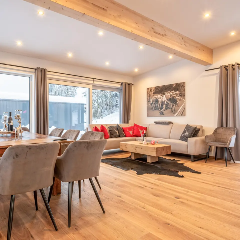 The living and dining room: a wooden floor, a timber beam, a corner sofa with red cushions and a dining table for ten; snow outside.