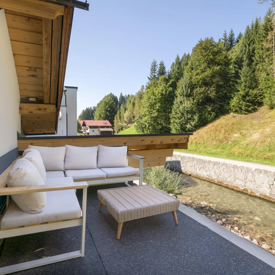 The terrace with a lounge sofa and a low table, the stream running right alongside and a stone wall on the far bank.